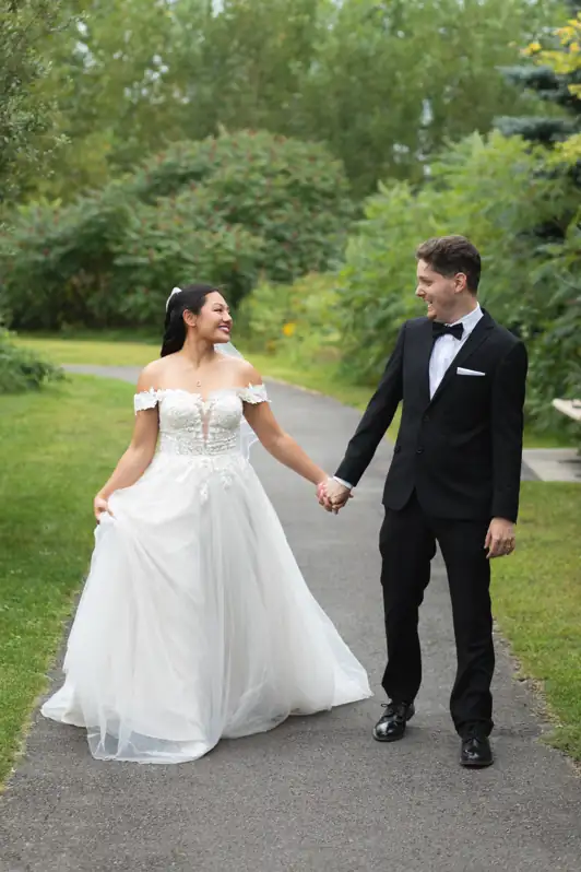 Bride and groom walking hand-in-hand and smiling on a lush, tree-lined path in Orléans, Ontario, during a summer wedding.