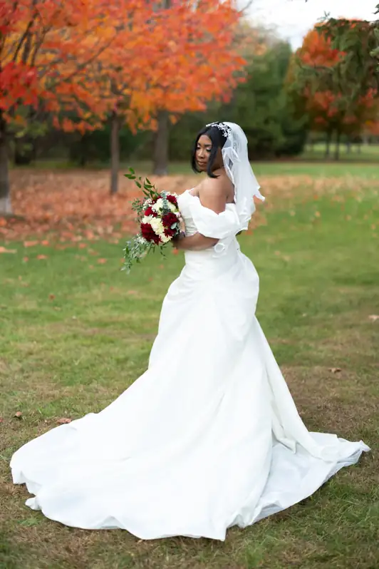 Bride in a white wedding dress looking down at her bouquet in an autumn field filled with vibrant fall foliage in Ottawa