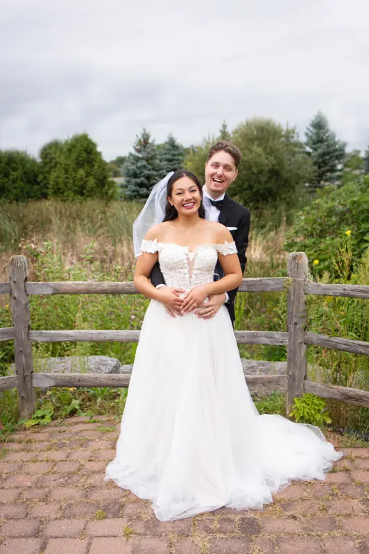 A groom hugging his bride from behind with both of them wearing wide, joyful smiles during their outdoor wedding portraits in Ottawa.