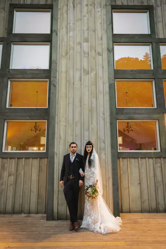 A bride and groom standing in a serious, editorial-style pose between two large windows outside their wedding reception venue.