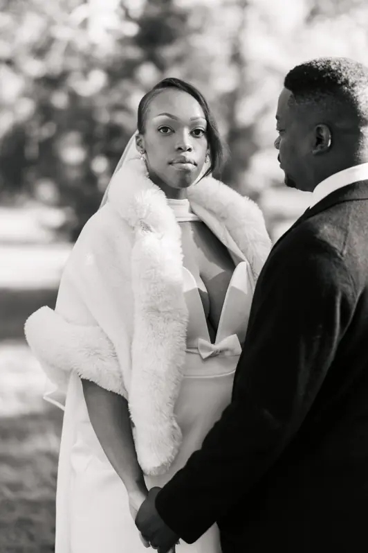 A close-up portrait of a bride looking pensively at the camera over her husband's shoulder while they hold hands tightly.