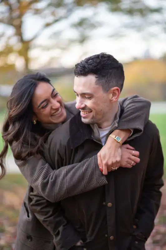 A joyful shot of a fiancée hugging her partner from behind in an Ottawa park; both are laughing candidly with a blurred background of green summer trees.