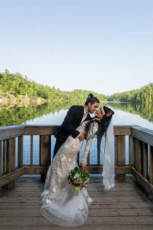 A groom tipping his wife backwards in a romantic dip pose on the lookout boards at Pink Lake in Gatineau Park, with the vibrant green water in the background.
