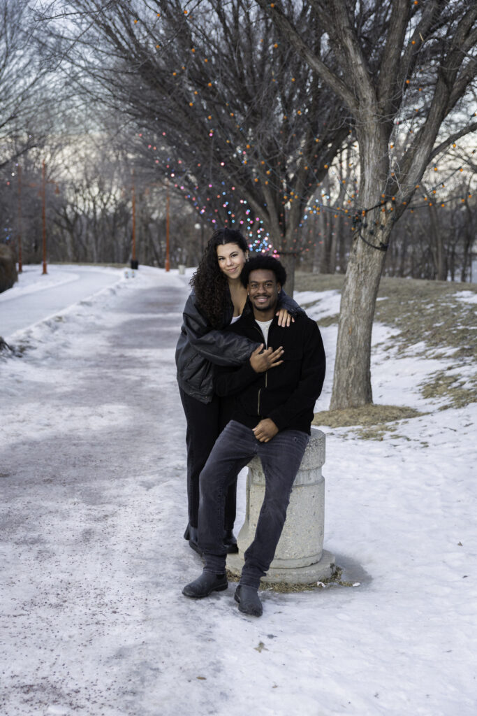 A couple sitting together in a snowy Ottawa park; the woman is wrapping her arms around her partner from behind, and both are smiling warmly at the camera.